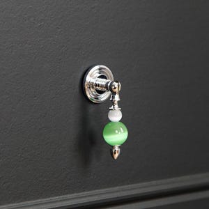 Pode incluir: Um puxador de gaveta em metal prateado com uma placa de base circular e uma pega vertical pendente. A pega &eacute; decorada com uma pequena conta esf&eacute;rica branca e uma conta esf&eacute;rica maior de cor verde-claro transl&uacute;cida, terminada com uma ponta met&aacute;lica.