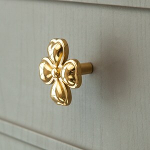 May include: Gold-toned flower-shaped cabinet knob with a rounded, slightly raised design. The knob is attached to a small, round, metal base.