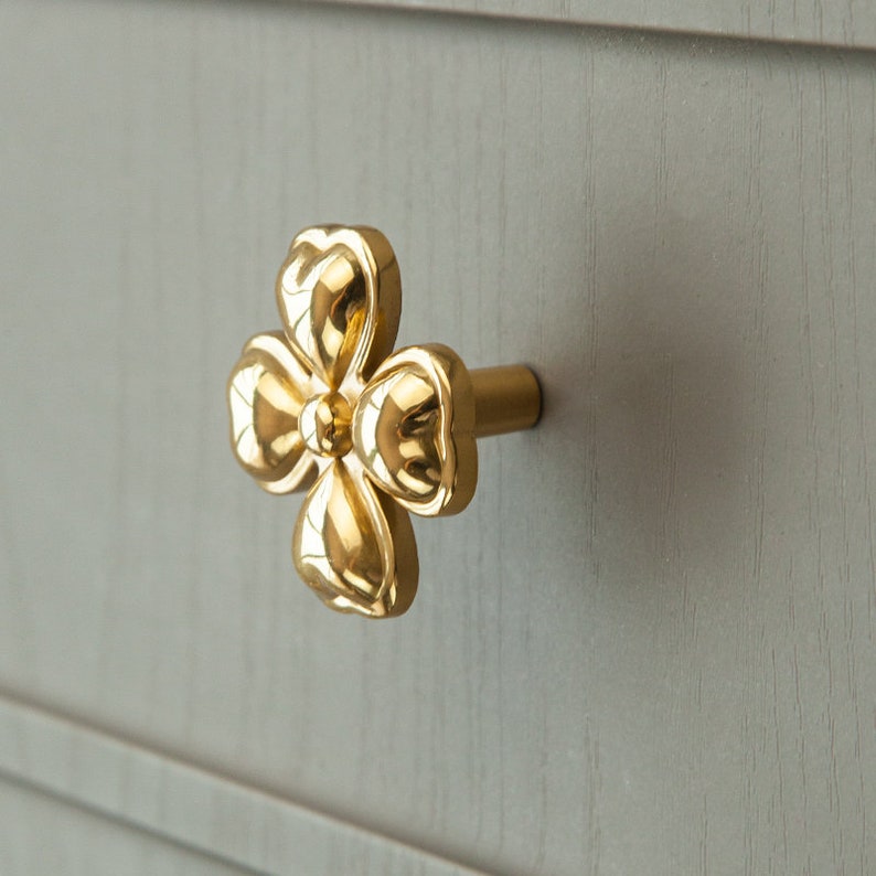 May include: Gold-toned flower-shaped cabinet knob with a rounded, slightly raised design. The knob is attached to a small, round, metal base.