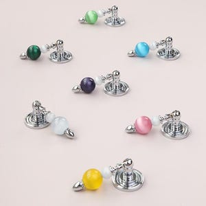 May include: Seven cabinet pulls with a circular silver-toned metal base, a short arm, a small white bead, and a larger spherical cat's eye stone in various colors including light green, dark green, blue, purple, white, pink, and yellow.