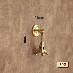 Peut inclure: Un bouton de meuble dor&eacute; avec un design de la Tour Eiffel. Le bouton mesure 24 mm de large et 67 mm de haut. Le poids est de 39g. Le bouton est mont&eacute; sur une surface marron.