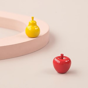 May include: A yellow pear-shaped object and a red apple-shaped object. The pear has a stem and is sitting on a pink, curved surface. The apple has a stem and is on a white surface. Both objects have a glossy finish.