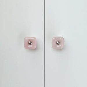 May include: Two square, pale pink cabinet knobs with rounded corners and a smooth, glossy finish. Each knob features a small, centered, circular silver-toned metal accent piece that sits flush within the middle of the pink square surface.