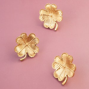 Peut inclure: Trois breloques tr&egrave;fles &agrave; quatre feuilles dor&eacute;s sur fond rose. Les breloques sont d&eacute;taill&eacute;es avec des lignes textur&eacute;es.