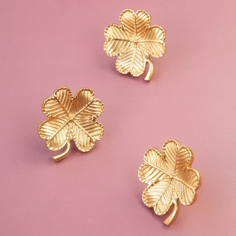 Peut inclure: Trois breloques tr&egrave;fles &agrave; quatre feuilles dor&eacute;s sur fond rose. Les breloques sont d&eacute;taill&eacute;es avec des lignes textur&eacute;es.