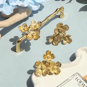 Puede incluir: Herrajes de armario de color dorado con un diseño floral. El herraje presenta una rama con delicados acentos florales.