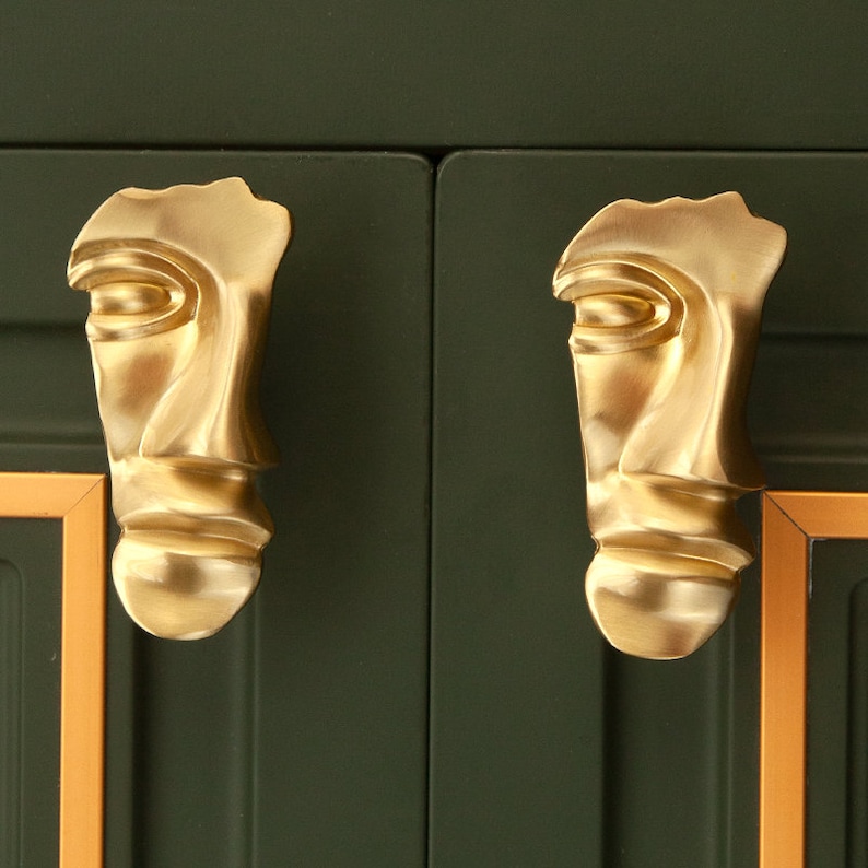 May include: Two gold-toned face-shaped cabinet knobs with a stylized design. The knobs are mounted on a dark green cabinet door.