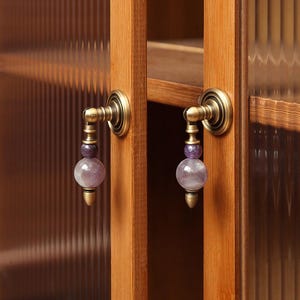 May include: Close-up of cabinet door hardware. The antique brass handles feature a decorative design with a purple amethyst gemstone and a conical base. The cabinet doors are made of wood and have a ribbed glass panel.