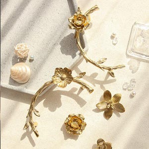 May include: Gold-colored floral branch-shaped cabinet pulls and knobs. The hardware features detailed flower and leaf designs. Decorative elements include a seashell, pearls, and a woven ball.