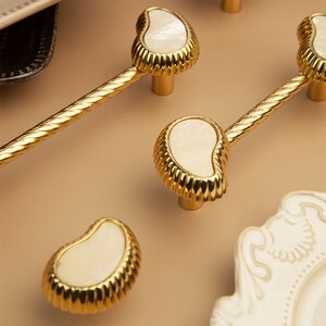 May include: Gold cabinet knobs with a pearlescent white shell inlay. The knobs have a rope-like design on the stem.