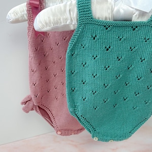 May include: Two knitted baby onesies, one in pink and one in green, hanging on a white hanger. The onesies have a lacy pattern and are made with soft yarn. The text "Knitting Pattern. ENGLISH" is visible in the image.