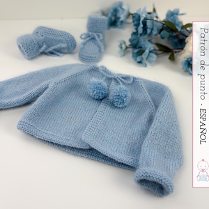 Puede incluir: Conjunto de bebé de punto azul claro, que incluye una rebeca con pompones y patucos a juego. El conjunto se exhibe sobre una superficie blanca con detalles florales azules. La rebeca tiene cuello redondo y mangas largas.