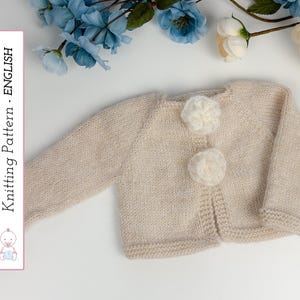 Puede incluir: Un cárdigan de bebé tejido de color beige claro con mangas largas y dos botones blancos en forma de flor. El cárdigan se muestra sobre una superficie blanca con flores azules y blancas en el fondo. El texto "Knitting Pattern - ENGLISH" es visible.