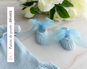 Op de afbeelding: Een paar lichtblauwe gebreide babyschoentjes met blauwe strikken. De schoentjes liggen op een wit oppervlak met een lichtblauwe gebreide babyjurk op de achtergrond. De tekst "Patrón de punto - ESPAÑOL" is zichtbaar op de jurk.