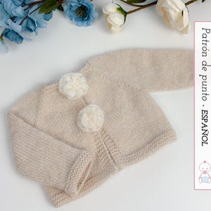 May include: A light beige knitted baby cardigan with two white flower-shaped buttons. The cardigan has long sleeves and a ribbed hem and cuffs. The text "Patrón de punto - ESPAÑOL" is visible on the right side of the image.