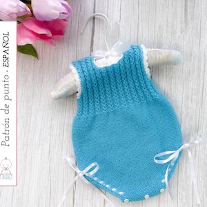May include: A light blue knitted baby romper with white trim and ribbons, displayed on a white hanger. The romper features a textured knit pattern and is adorned with white ribbons at the sides. The image also includes text in Spanish.