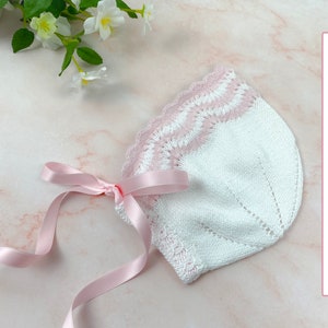 May include: A white and pink knitted baby bonnet with a scalloped edge and a pink satin ribbon tied around it. Knitting Pattern - ENGLISH.