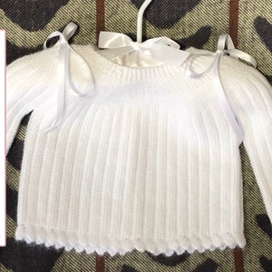 May include: White knitted baby sweater with long sleeves and white satin ribbon ties at the shoulders. The sweater has a ribbed texture and a scalloped hem.
