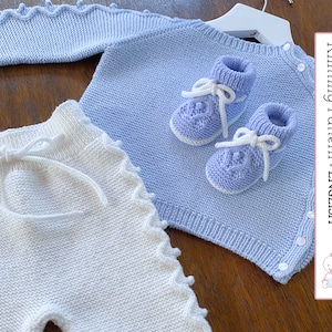 May include: A knitted baby outfit set in blue and white. The set includes a sweater, pants, and booties. The sweater has a button closure and the pants have a drawstring. The booties have a white trim and laces.