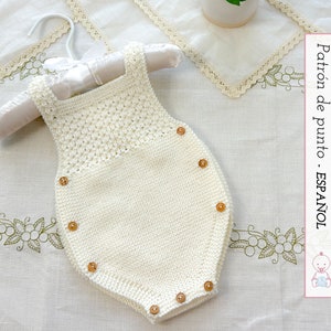 Puede incluir: Un body de bebé de crochet blanco con botones de madera. El body está colgado de una percha blanca y se encuentra sobre un fondo blanco con un patrón floral.
