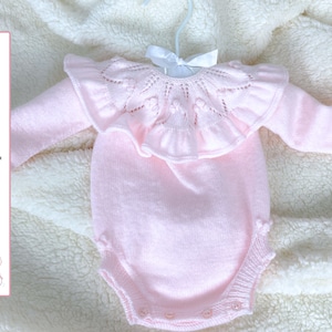 Könnte beinhalten: Ein rosa gestrickter Baby-Strampler mit einem Spitzenkragen mit Rüschen. Der Strampler hat lange Ärmel und einen Druckknopfverschluss am unteren Ende. Der Text "Patrón de punto - ESPAÑOL" ist auf der linken Seite des Bildes sichtbar.
