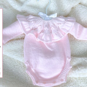 Puede incluir: Un pelele de bebé de punto rosa con un cuello de encaje con volantes y mangas largas. El pelele está sobre una superficie blanca y esponjosa.