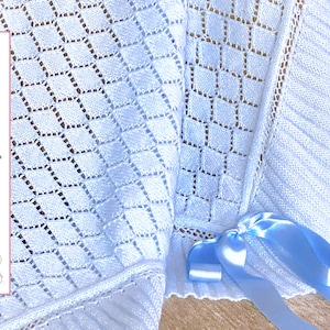 Puede incluir: Una manta de bebé de crochet blanco con un delicado patrón de diamantes. La manta está atada con una cinta de satén azul claro. El texto "Patrón de punto - ESPAÑOL" es visible en la imagen.