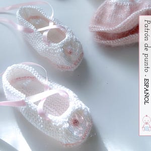 May include: Hand-knitted white baby booties with pink ribbon ties and accents. The booties have a delicate, textured appearance, with a small pink embellishment on the toe. A pair of pink knitted socks are also visible.
