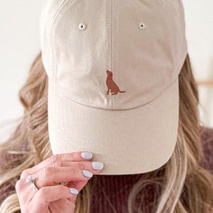 Könnte beinhalten: Eine beige Baseballkappe mit einer braunen gestickten Hundesilhouette auf der Vorderseite. Die Kappe wird von einer Person mit langen blonden Haaren gehalten.