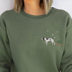 May include: Green sweatshirt featuring an embroidered design of a white dog with black spots pulling a small Christmas tree on a sled. The design is on the front of the sweatshirt.
