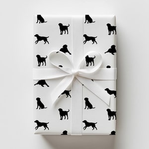 May include: Wrapped gift with white paper featuring a repeating pattern of black dog silhouettes. A white satin ribbon is tied in a bow around the package. The gift is presented on a white background.