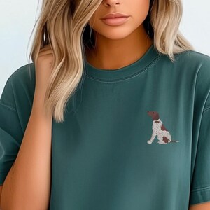 May include: A blue spruce green t-shirt with a small embroidered image of a brown and white dog. The text "COLOR: BLUE SPRUCE" is visible at the bottom of the image.