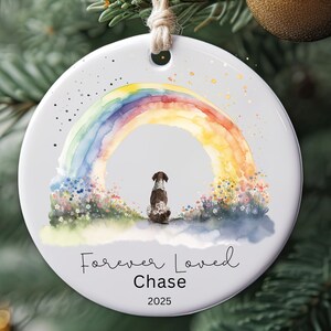 Könnte beinhalten: Runder, weißer Keramik-Weihnachtsschmuck mit einem Aquarell-Regenbogen und einem Hund. Der Text "Forever Loved Chase 2025" ist abgebildet. Eine Juteschnur ist zum Aufhängen befestigt. Der Hintergrund umfasst einen Weihnachtsbaum und eine goldene Kugel.
