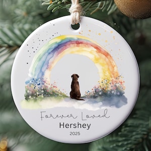 May include: Round white Christmas ornament with a watercolor rainbow design and a brown dog silhouette. The text "Forever Loved Hershey 2025" is written below the dog and rainbow. The ornament hangs from a natural twine.