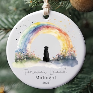 May include: Round white ceramic Christmas ornament featuring a watercolor rainbow and a black dog silhouette. The text "Forever Loved Midnight 2025" is written below the image. A jute string is attached for hanging.