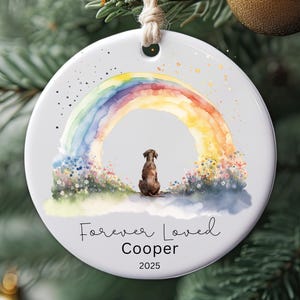 May include: A circular white ceramic Christmas ornament featuring a watercolour rainbow and a brown dog. The text "Forever Loved Cooper 2025" is written below the dog and rainbow. The ornament is suspended by natural twine.