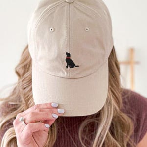 Könnte beinhalten: Beige Baseball Cap mit einer schwarzen Hunde-Silhouette und rotem Halsband auf der Vorderseite. Die Kappe hat einen gebogenen Schirm und einen verstellbaren Riemen auf der Rückseite.