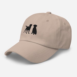 May include: A beige baseball cap with an embroidered design of two black silhouettes of dogs. One dog is standing and the other is sitting.