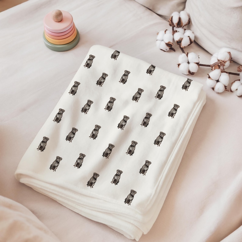 Pu&ograve; includere: Una coperta per bambini bianca piegata con un motivo ripetuto di piccoli cani marroni seduti. La coperta &egrave; realizzata in materiale morbido ed &egrave; piegata con cura. Un giocattolo impilabile in legno e rametti di cotone sono sullo sfondo.