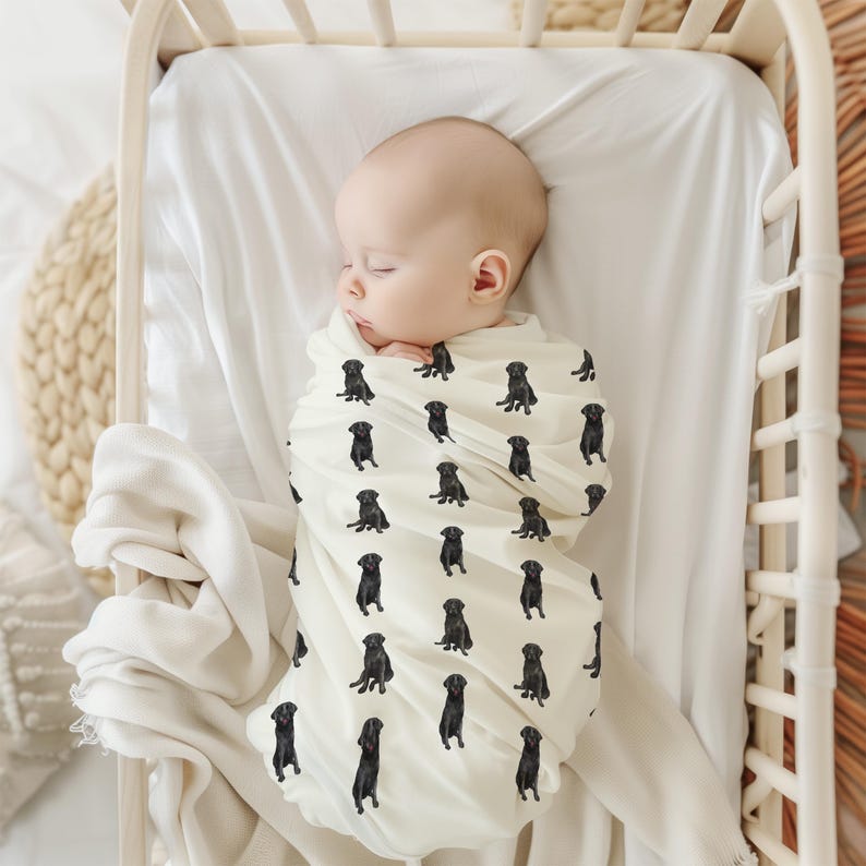 May include: A baby swaddled in a white muslin swaddle blanket with a black Labrador Retriever print. The baby is sleeping peacefully in a white crib.