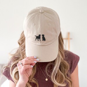 May include: A beige baseball cap with two black silhouette dogs facing each other on the front.