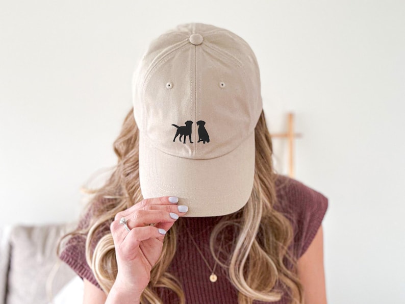 May include: A beige baseball cap with two black silhouette dogs facing each other on the front.