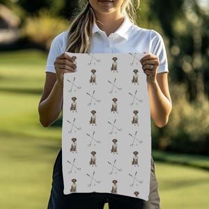 May include: A light gray towel featuring a repeating pattern of a brown dog and crossed golf clubs. The dog is sitting, and the golf clubs are white with a black grip. The towel is held by a person.