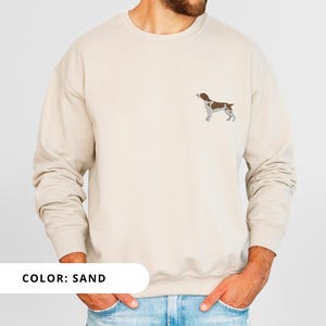May include: A sand colored sweatshirt with a brown and white dog silhouette embroidered on the front. The text "COLOR: SAND" is visible at the bottom of the image.