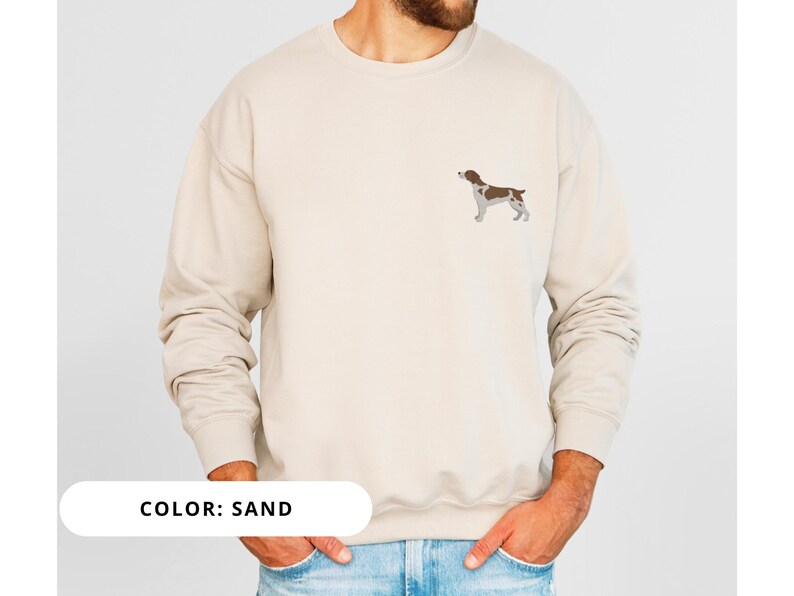 May include: A sand colored sweatshirt with a brown and white dog silhouette embroidered on the front. The text "COLOR: SAND" is visible at the bottom of the image.