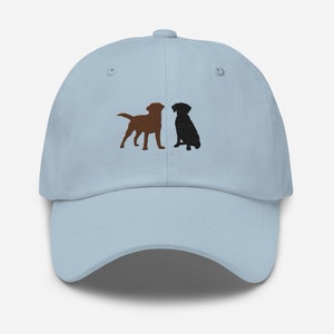 May include: A light blue baseball cap featuring an embroidered design of two labrador retrievers, one brown and one black, facing each other.