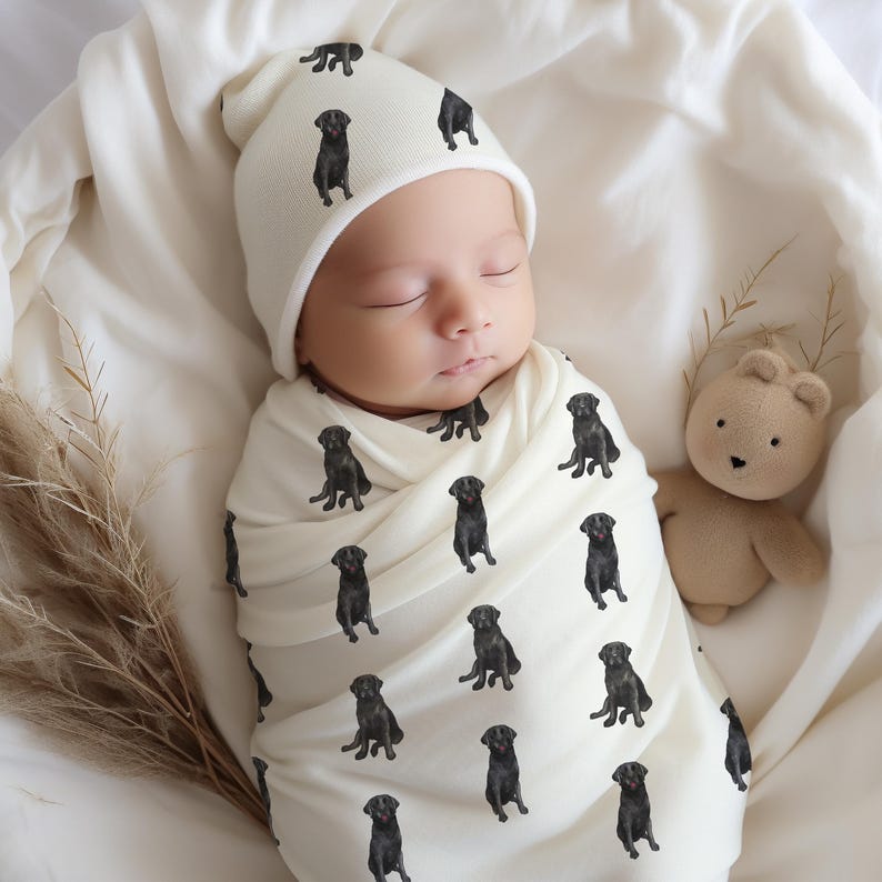 May include: A white swaddle blanket with a black Labrador Retriever print. A baby is sleeping in the swaddle, wearing a white beanie with a black Labrador Retriever print.