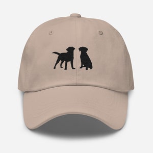 May include: A beige baseball cap with black embroidered silhouettes of two Labrador Retrievers facing each other.