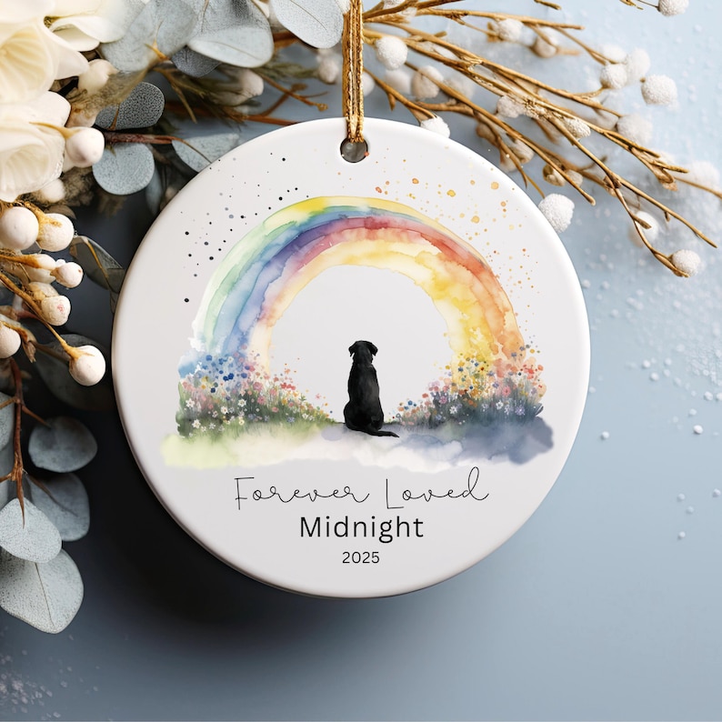 May include: A round white ceramic ornament featuring a watercolor rainbow and a black dog silhouette. The text "Forever Loved Midnight 2025" is written below the image. A gold string is attached for hanging.