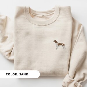 May include: A sand-colored sweatshirt with a brown and white dog illustration on the front. The text "COLOR: SAND" is printed below the illustration.